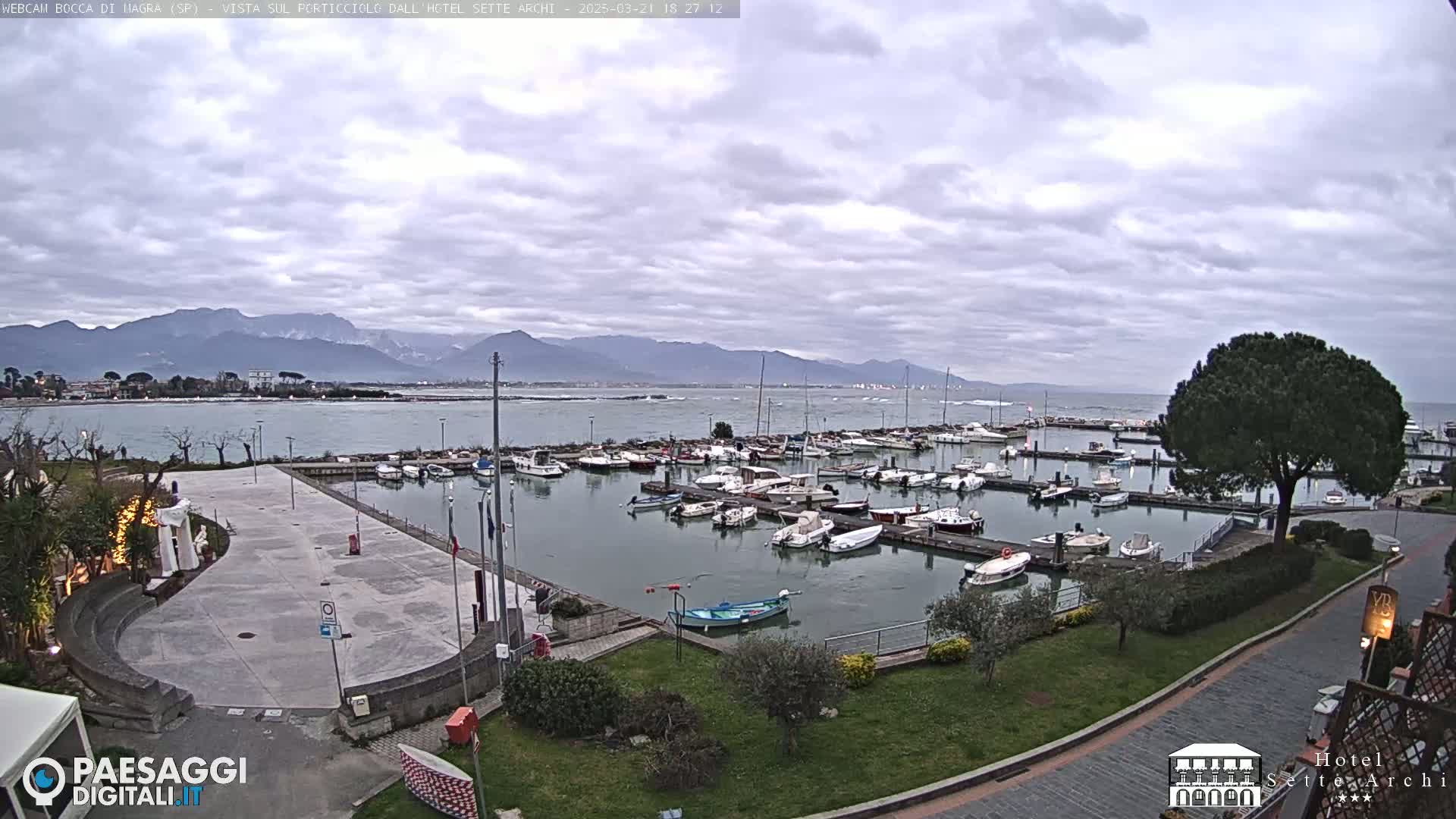 A harbor filled with numerous boats is visible under a cloudy sky, with mountains in the distance.