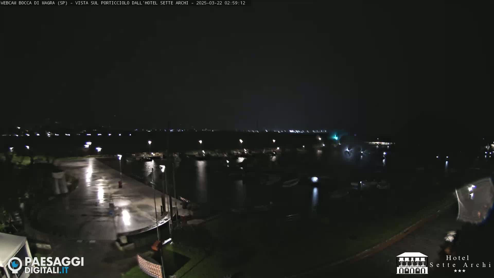 A nighttime view of a harbor with boats and a wet, empty paved area, under a dark sky.