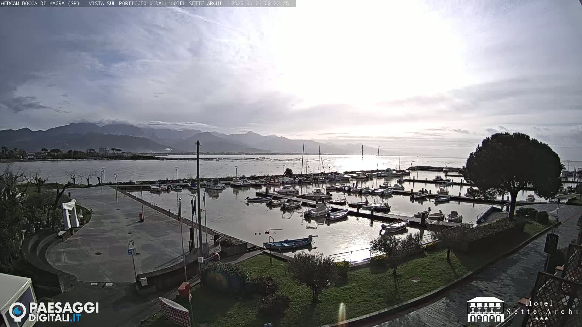 A mostly cloudy day reveals a marina filled with boats, situated next to a paved area and greenery, with mountains in the background.