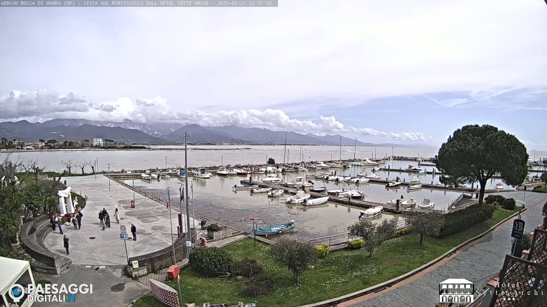 A partly cloudy day overlooks a marina filled with numerous boats, a paved plaza with several people, and distant mountains.