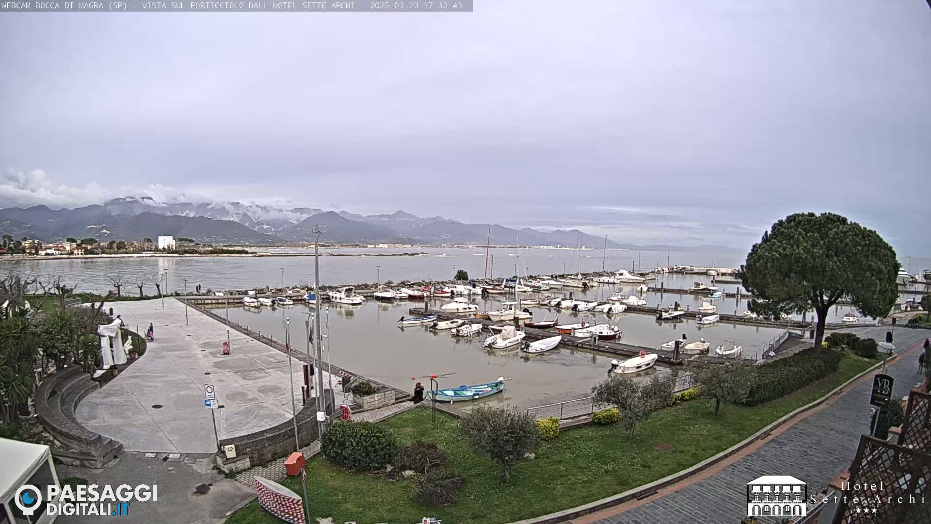 A harbor filled with numerous boats is situated near a paved plaza, all overlooking a calm body of water with snow-capped mountains in the distance under an overcast sky.