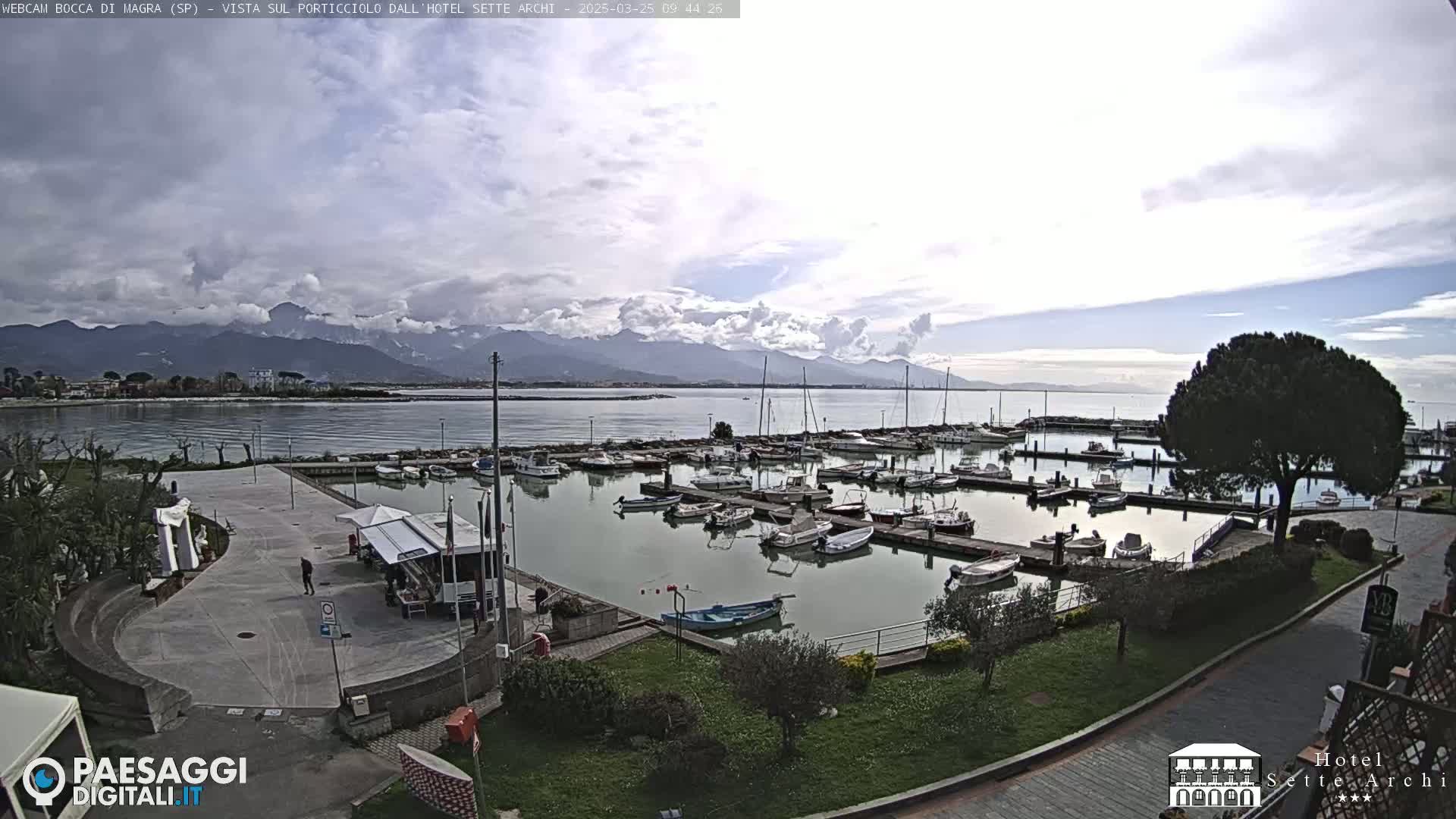 A marina filled with numerous boats is situated near a paved area, with mountains visible in the background under a partly cloudy sky.