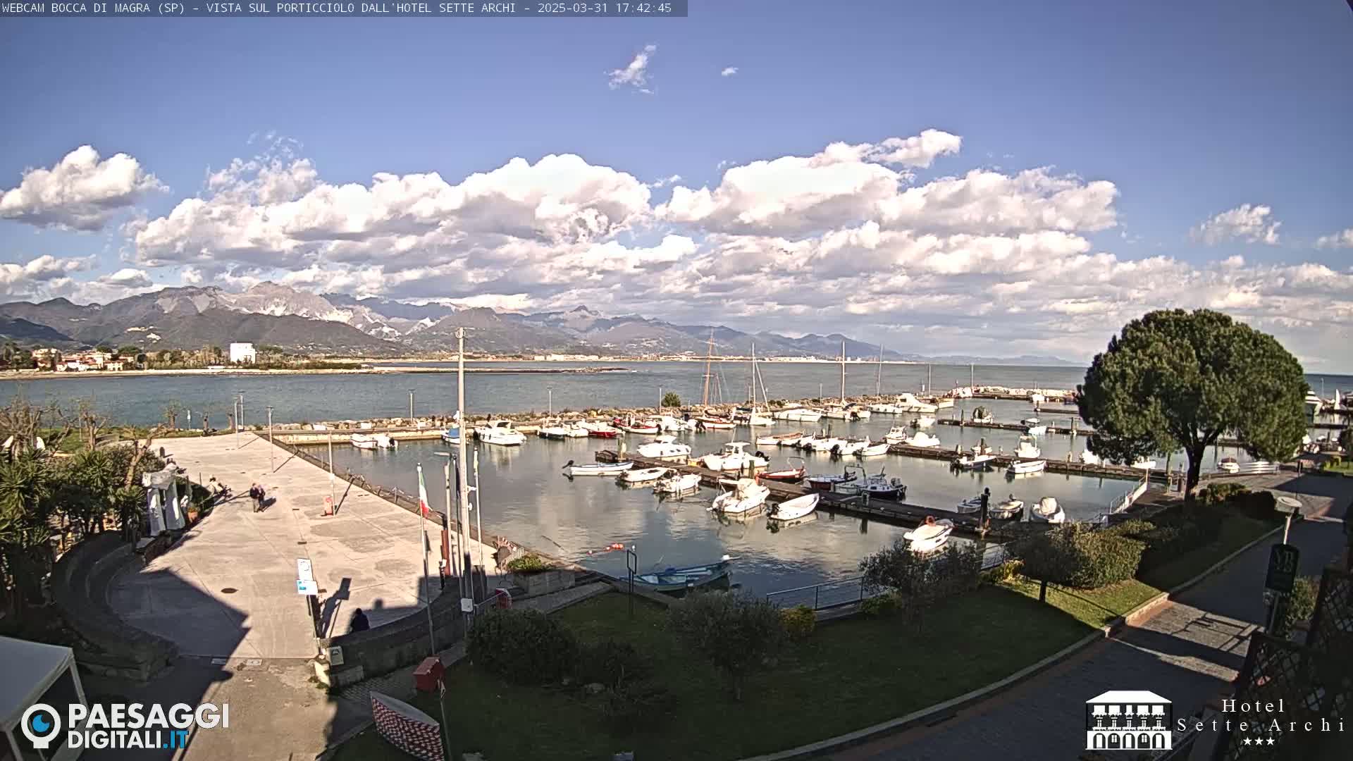 A partly sunny day shows a marina filled with boats, a paved area, and mountains in the distance.