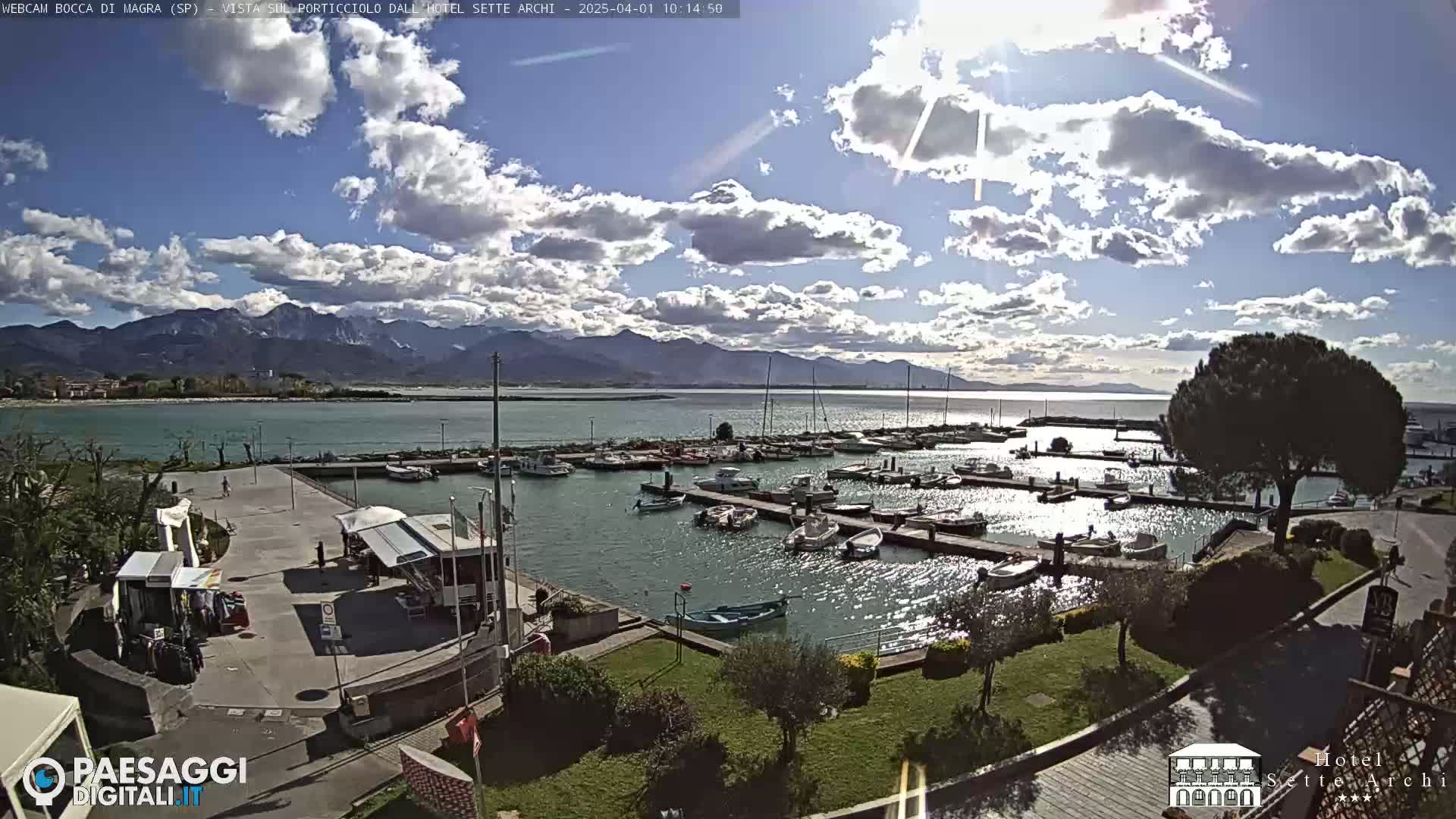 A sunny day with partly cloudy skies overlooks a harbor filled with boats, a paved area, and green landscaping, all set against a backdrop of mountains.
