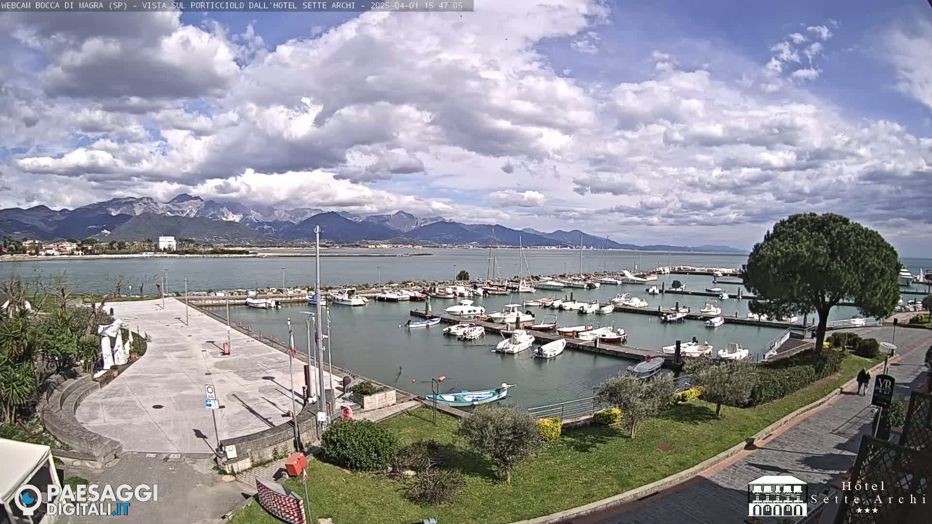 A partly cloudy day shows a marina filled with boats, a paved plaza, and mountains in the distance.