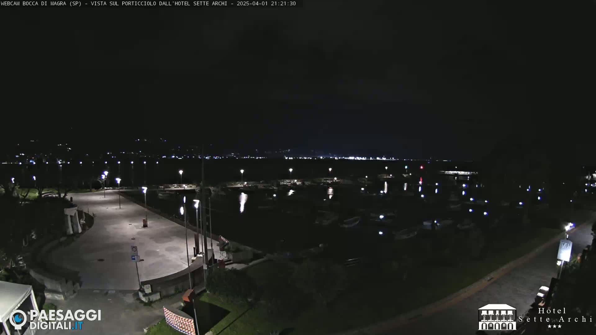 A nighttime view of a marina filled with boats, a paved waterfront area, and dimly lit surrounding landscape.
