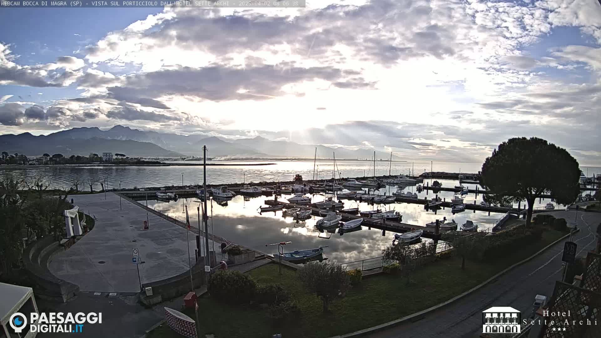 A marina filled with boats is situated near a calm body of water, under a partly cloudy sky, with mountains visible in the distance.