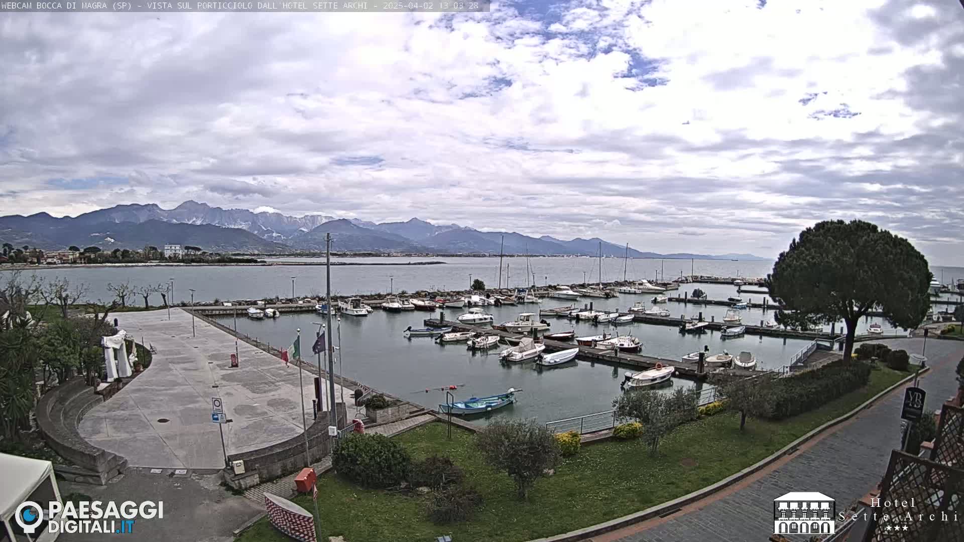 A marina filled with boats is situated near a paved plaza, with mountains visible in the distance under a partly cloudy sky.