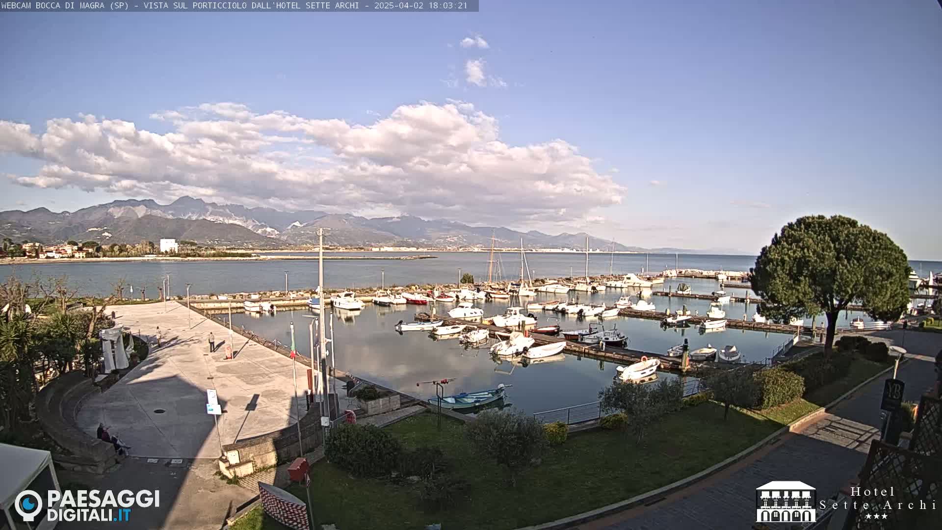 A marina filled with numerous boats is situated near a calm body of water, under a partly cloudy sky, with mountains visible in the distance.