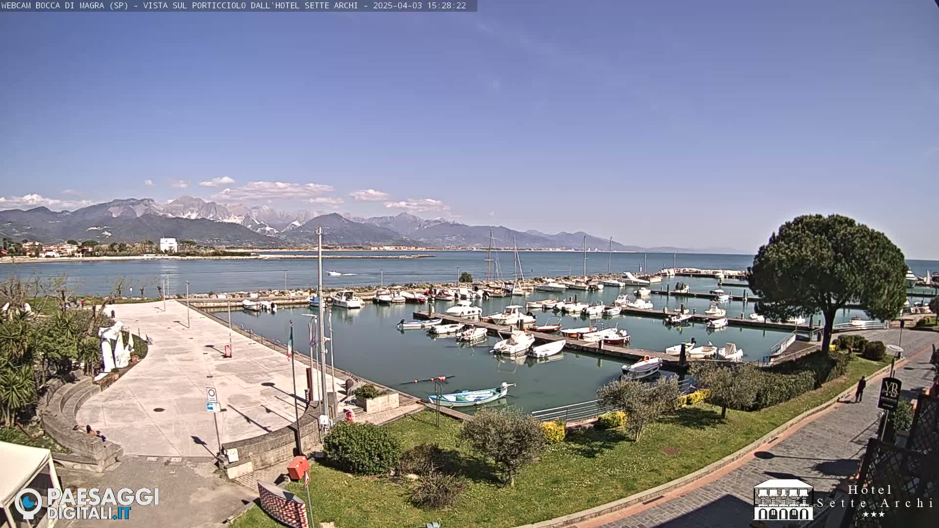 A marina filled with numerous boats is situated on calm water, with mountains in the background under a clear, sunny sky.