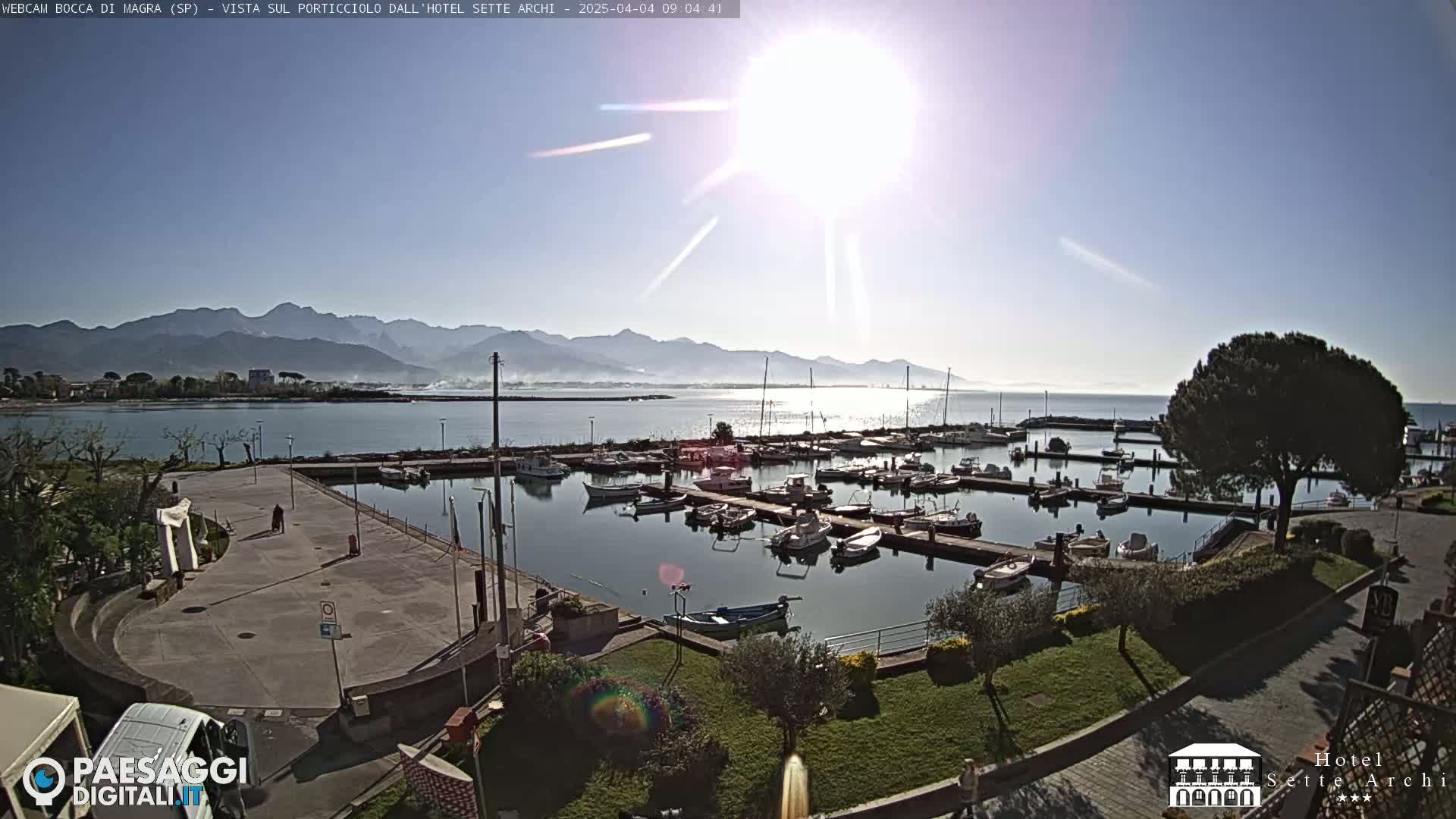 A sunny day shows a harbor filled with numerous boats, a paved area, and a distant mountain range under a mostly clear sky.