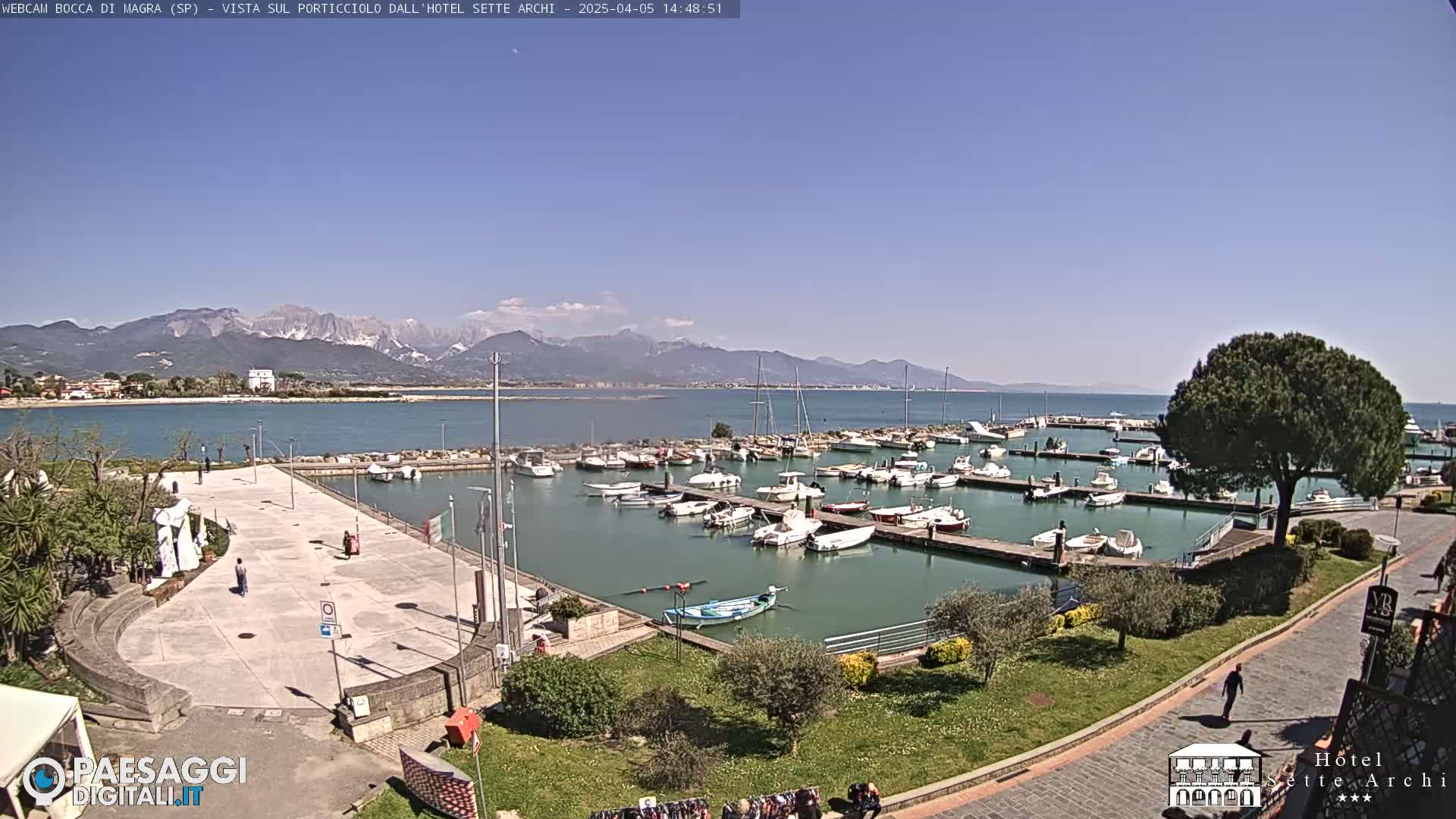 A harbor filled with boats sits next to a paved plaza, all under a clear blue sky and overlooking mountains in the distance.