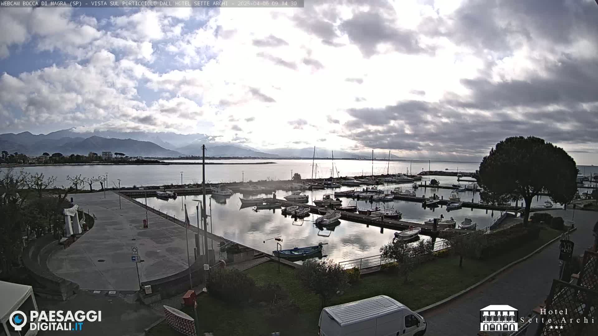 A marina filled with boats is seen under a cloudy sky, with mountains visible in the distance.