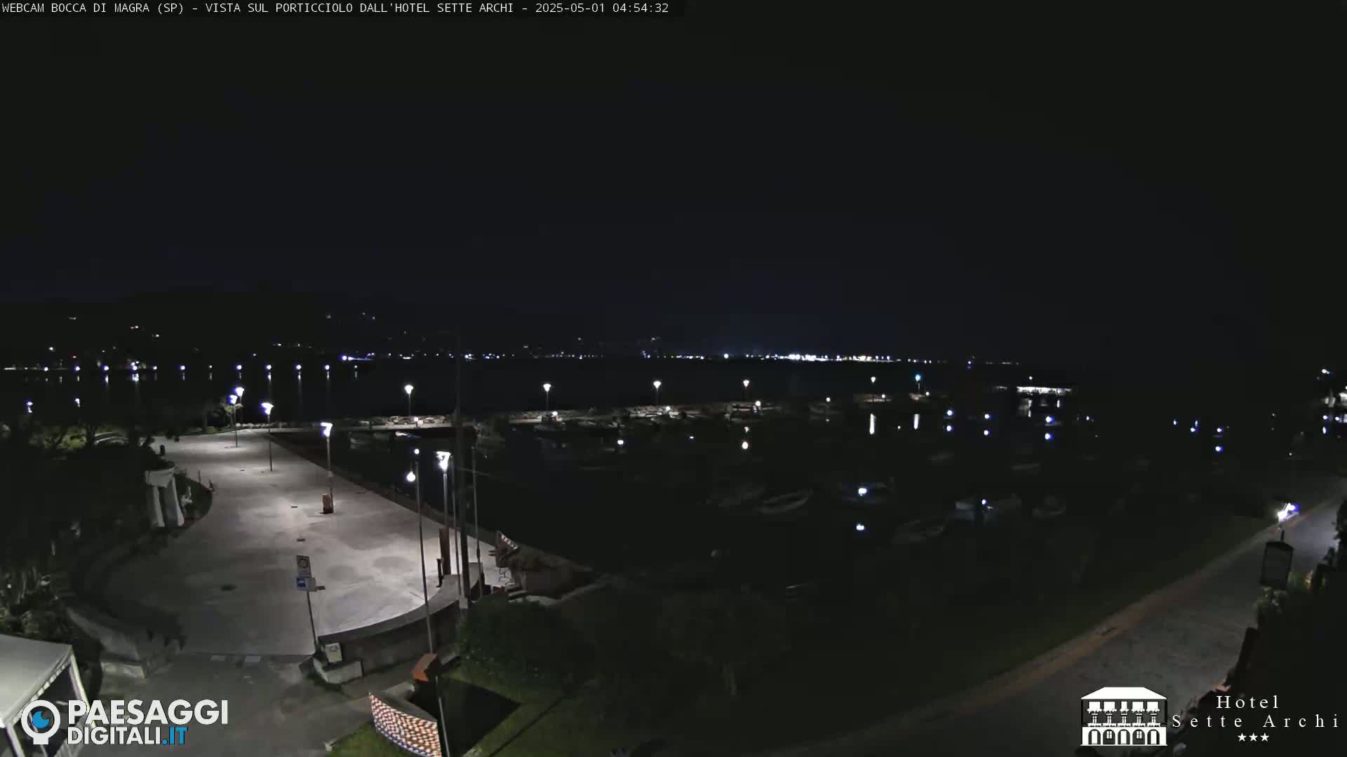 A nighttime view of a marina and paved waterfront area, illuminated by streetlights, with calm water reflecting the lights.
