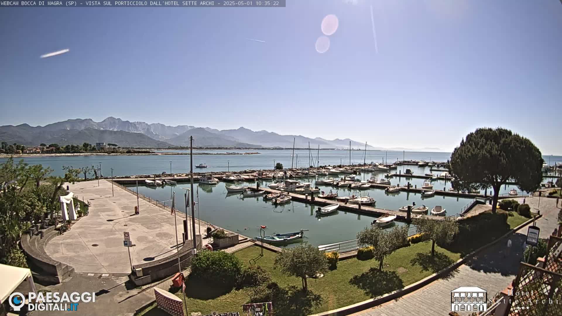 A sunny harbor filled with numerous boats is situated near a paved plaza and green landscaping, with mountains visible in the distance.