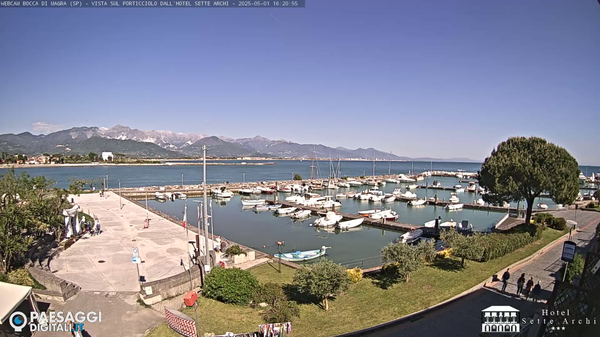 A marina filled with numerous boats is situated by a calm body of water, backed by mountains under a clear blue sky.