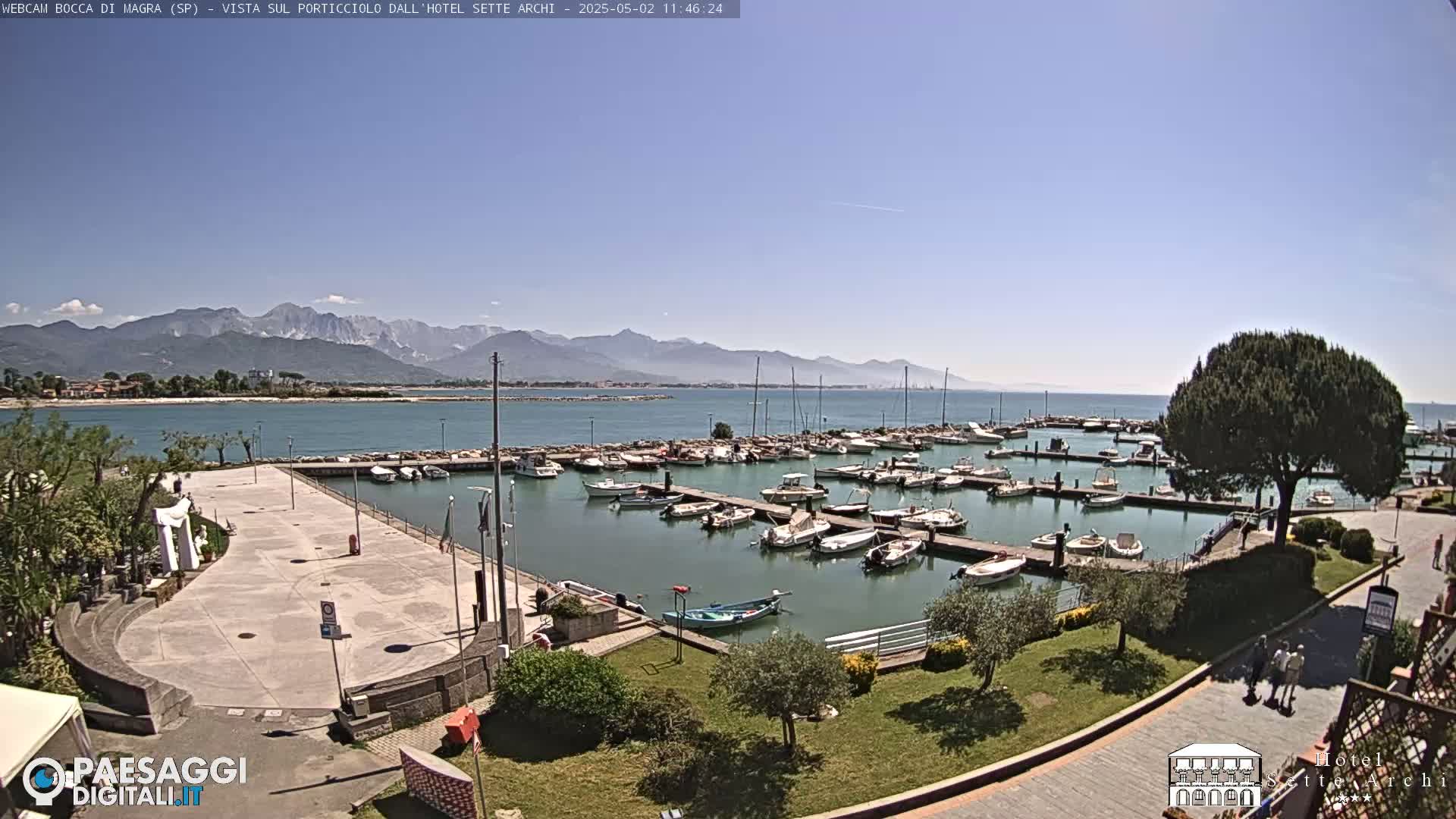 A marina filled with boats is situated by a calm body of water, backed by a sandy shore, under a clear blue sky and mountains in the distance.