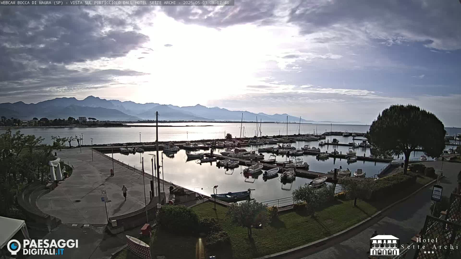 A marina filled with boats is situated near a calm body of water, with mountains in the background under a partly cloudy sky.