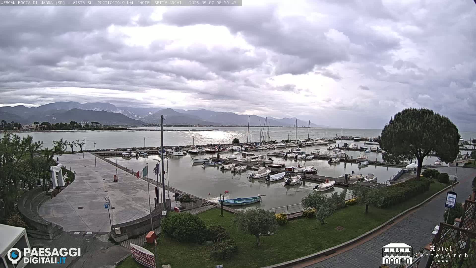 A marina filled with numerous boats is situated near a paved plaza and lush green landscaping, all under a cloudy sky and backed by distant mountains.
