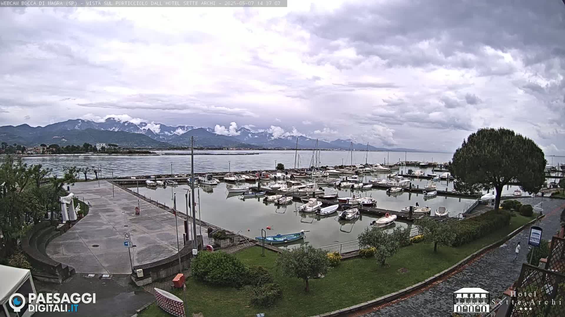 A marina filled with boats is situated near a paved plaza and grassy area, overlooking a calm body of water and distant mountains under a cloudy sky.