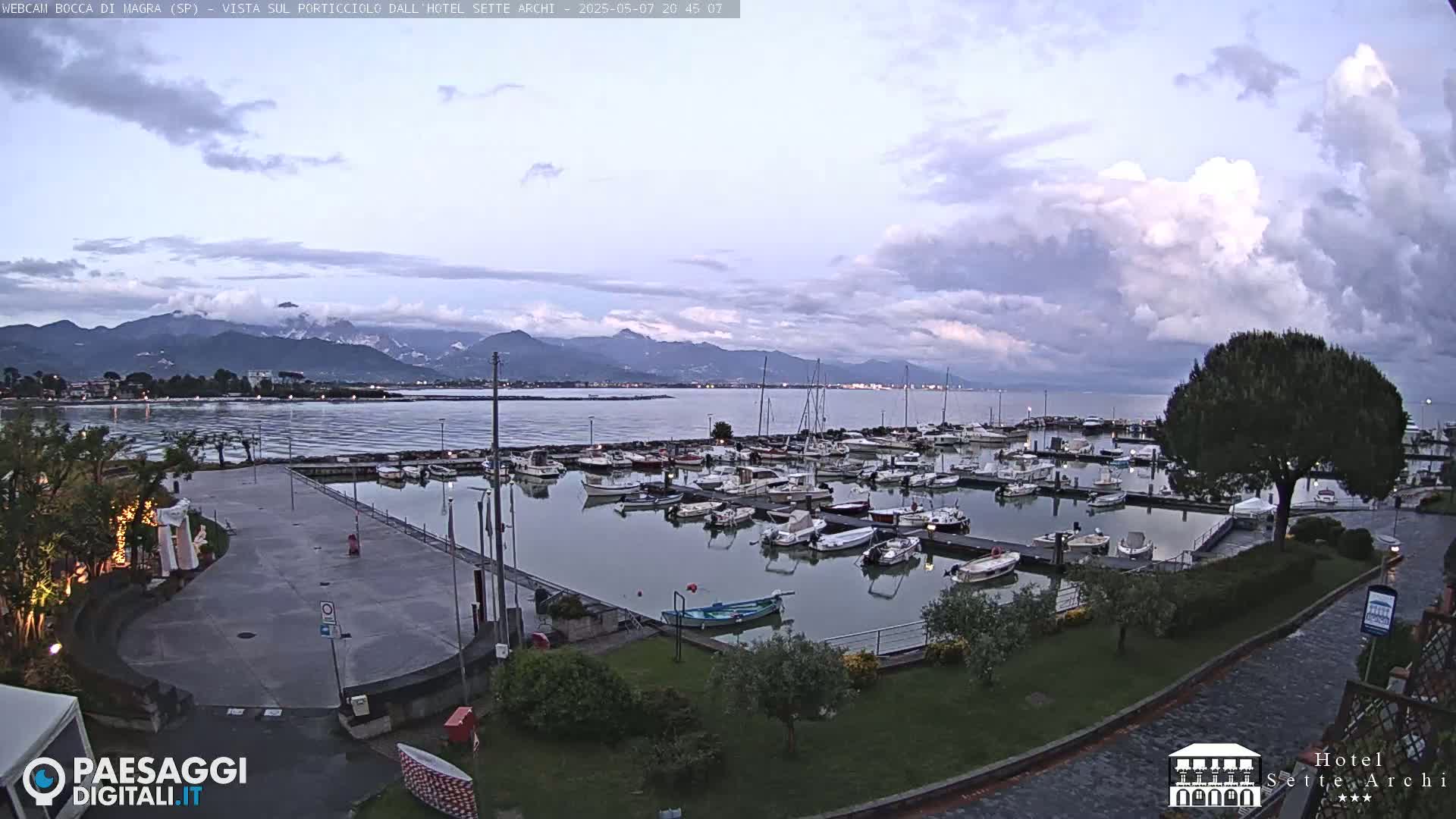 A harbor filled with boats is visible under a partly cloudy evening sky, with mountains in the distance.