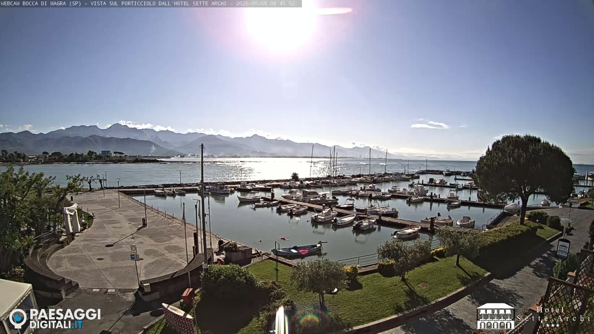 A sunny day reveals a marina filled with boats, a paved area, and lush green landscaping, all set against a backdrop of calm water and distant mountains.