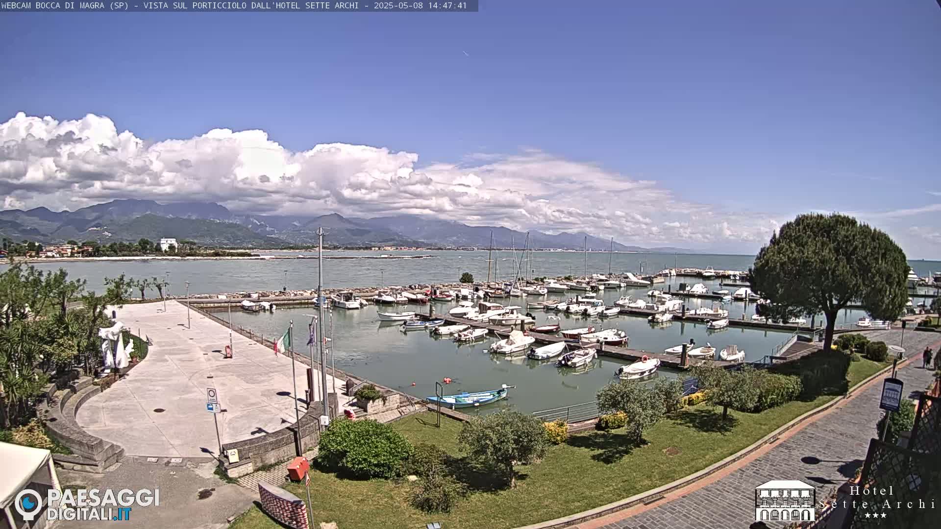 A partly cloudy day reveals a harbor filled with numerous boats, a paved plaza, and lush green landscaping leading to a calm body of water with mountains in the distance.