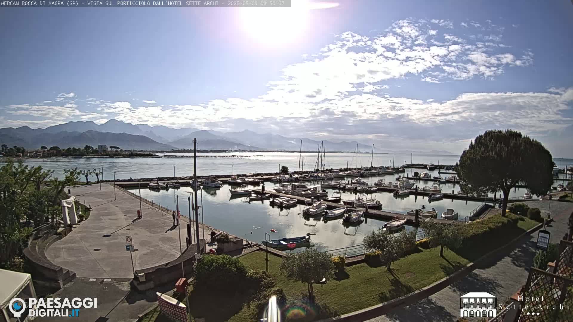 A partly cloudy day reveals a marina filled with boats, situated near a paved area and lush green landscaping, with mountains visible in the distance.