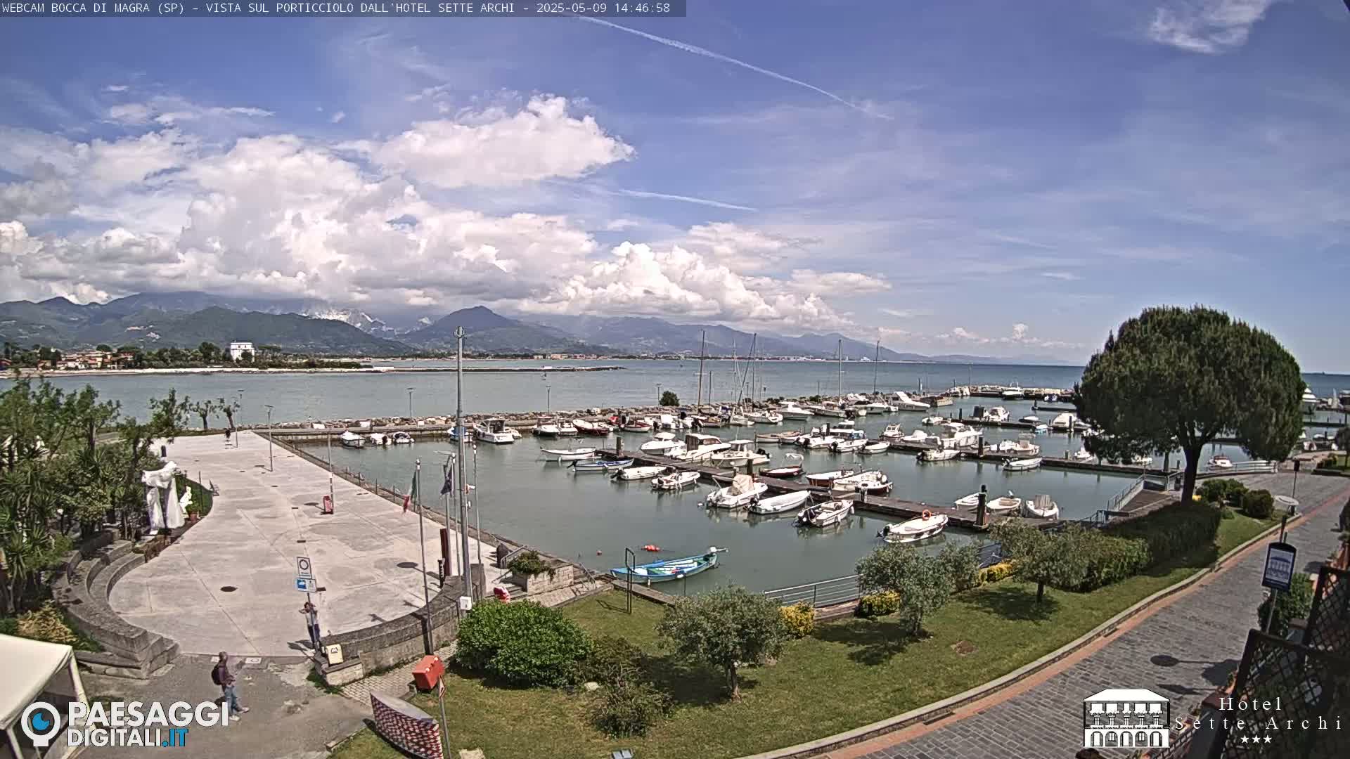 A marina filled with numerous boats is situated beside a paved plaza and green landscaping, all overlooking a calm body of water with mountains in the distance under a partly cloudy sky.