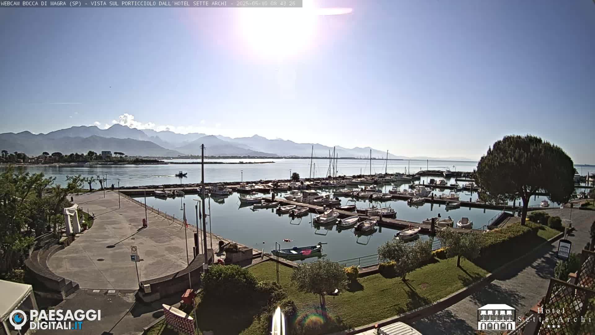 A sunny day overlooking a harbor filled with boats, a paved waterfront area, and mountains in the distance.