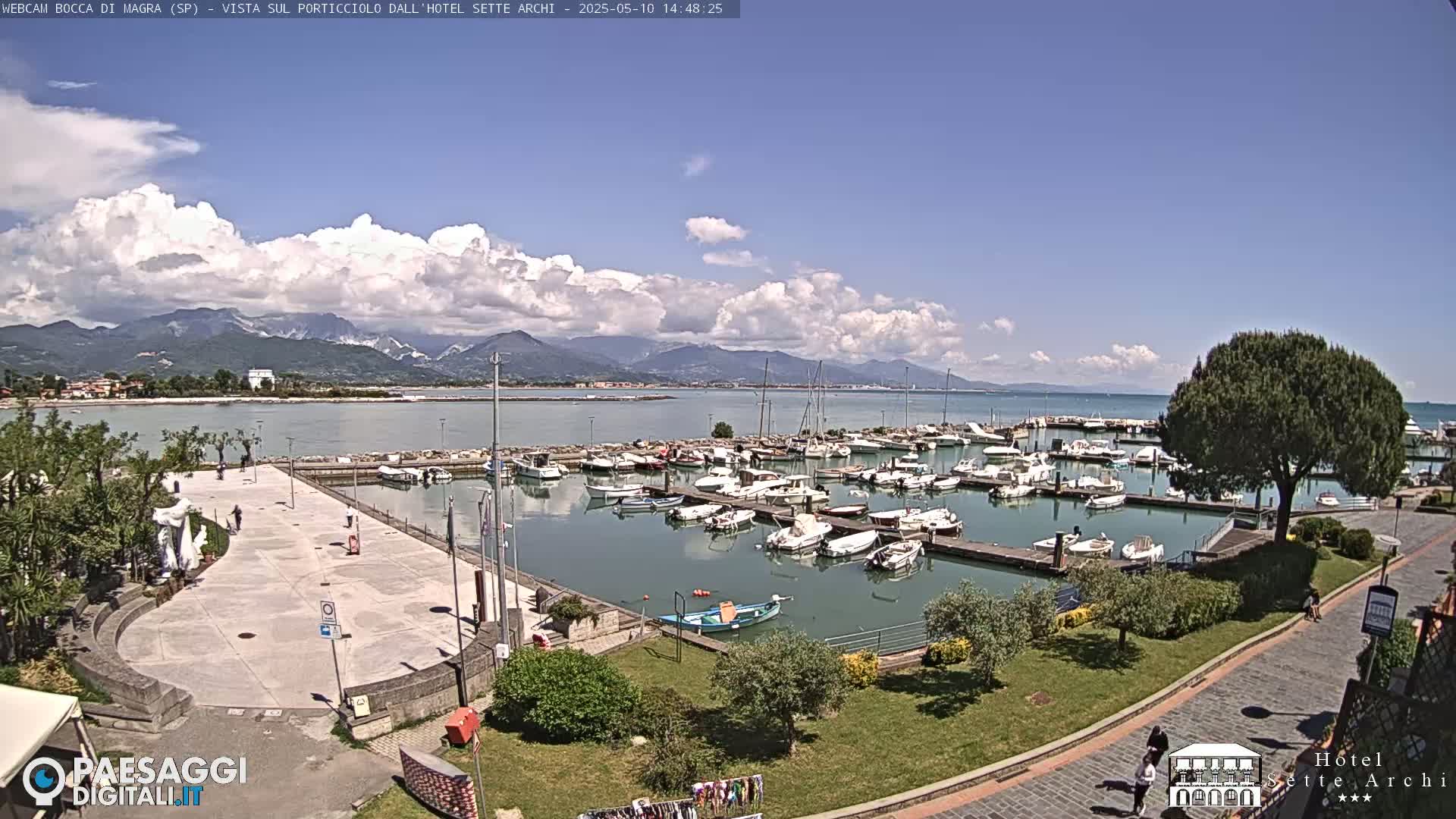 A marina filled with boats is situated on a calm body of water, with a paved plaza and green space nearby, all under a partly cloudy sky and mountains in the distance.