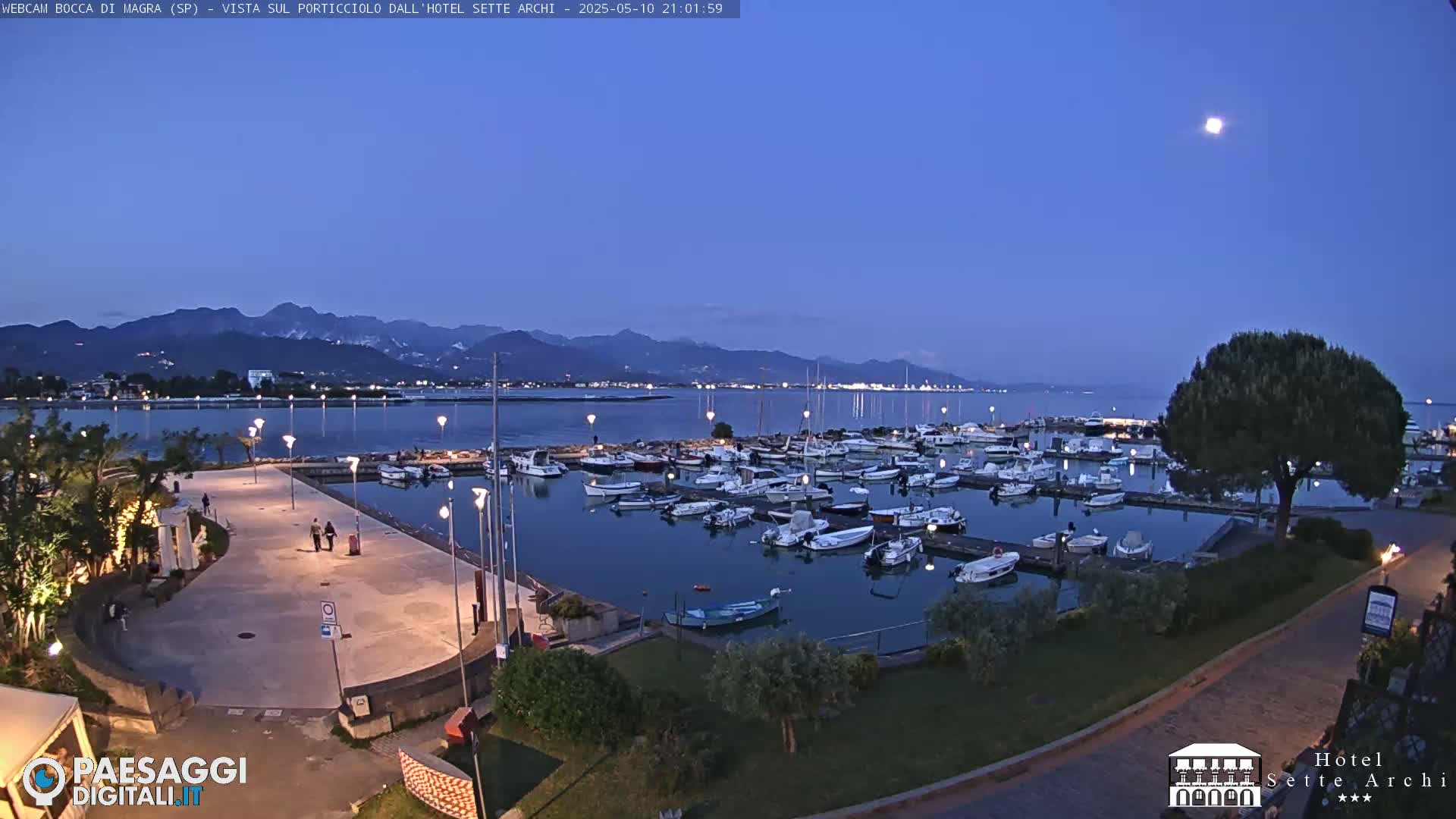 A marina filled with boats is situated along a paved waterfront path under a twilight sky, with mountains visible in the distance.