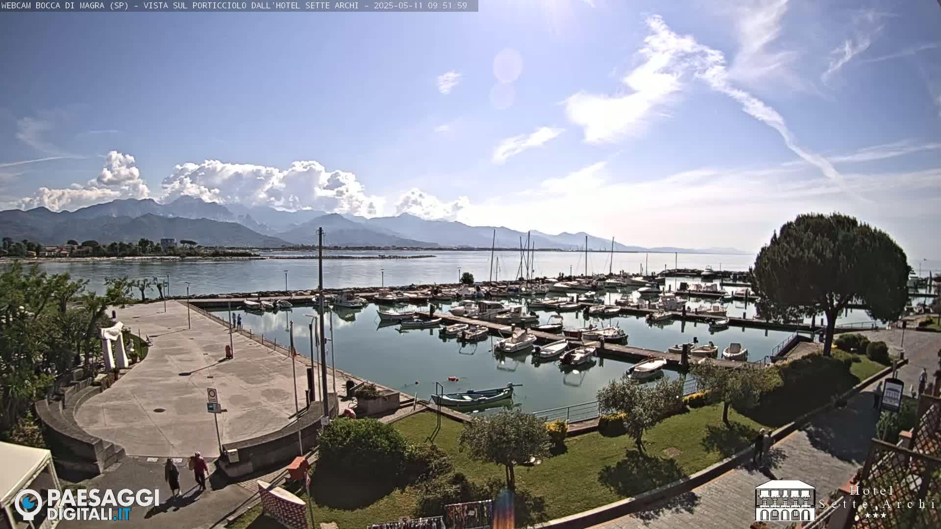 A marina filled with numerous boats is situated near a paved walkway and green landscaping, all under a partly cloudy sky with mountains in the background.