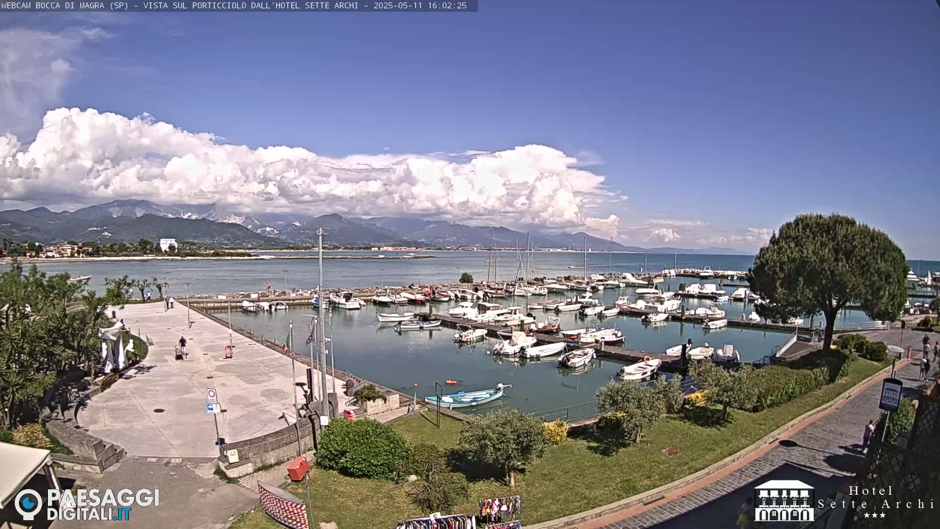 A harbor filled with numerous boats is situated near a paved plaza, overlooking a calm body of water with mountains in the background under a partly cloudy sky.