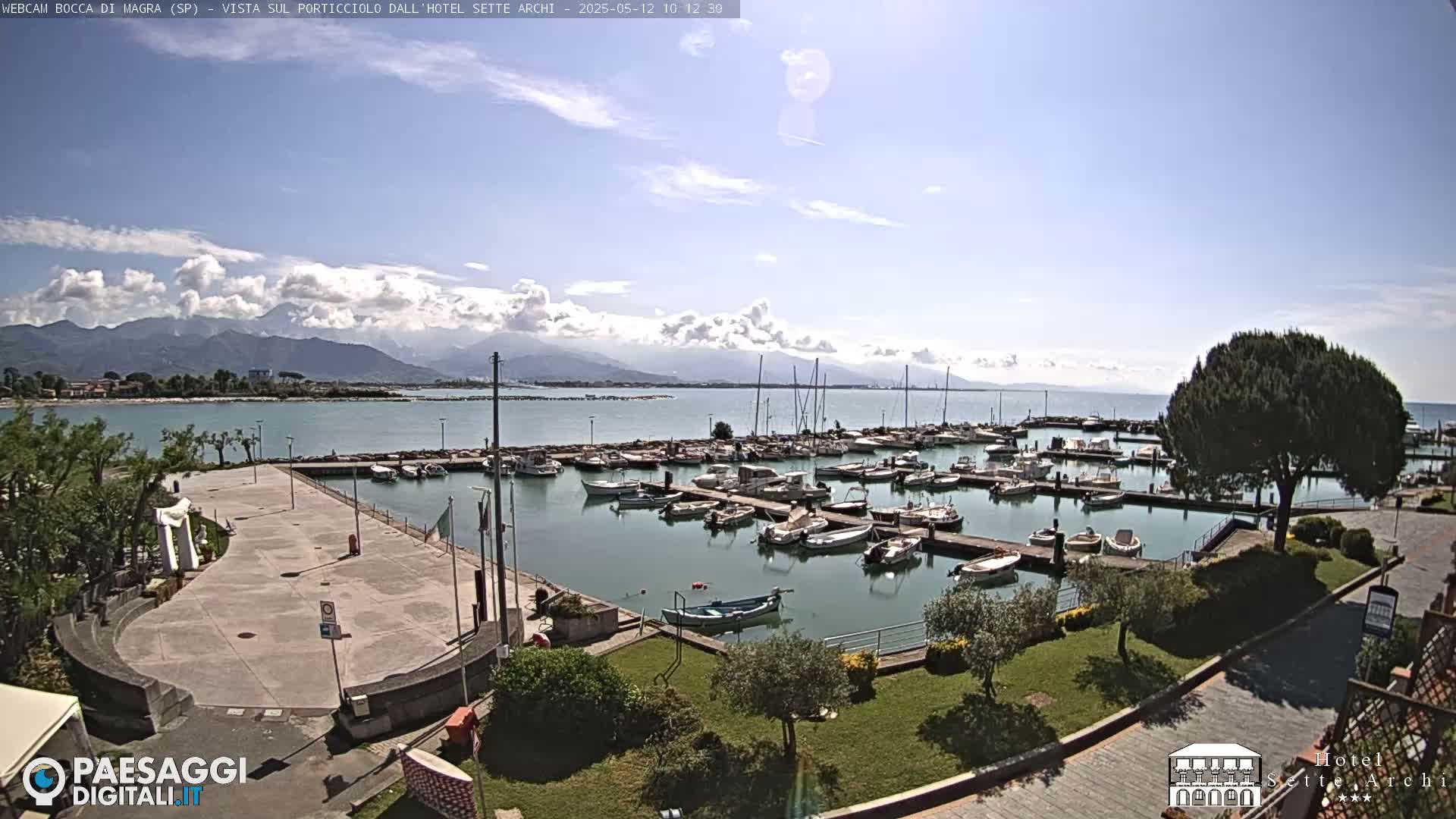 A marina filled with boats is situated on a calm body of water under a partly cloudy sky, with mountains visible in the distance.