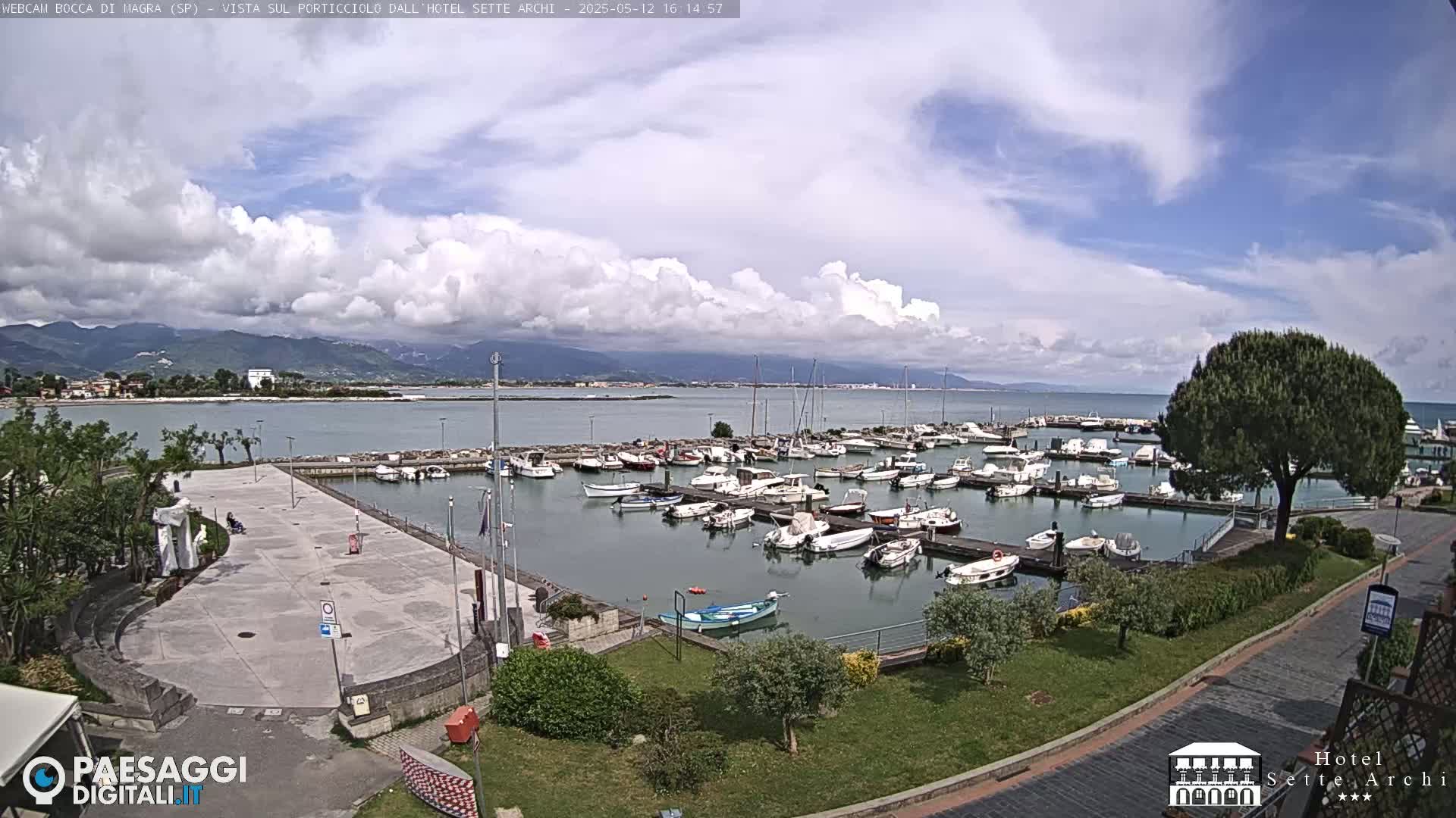 A partly cloudy day reveals a marina filled with boats, a paved plaza, and green landscaping, all overlooking a calm body of water and distant mountains.