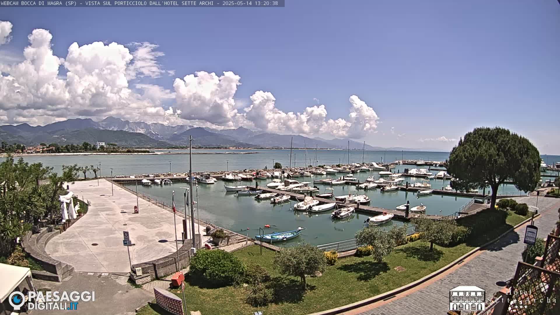 A partly cloudy day shows a marina filled with boats, a paved plaza, and green landscaping, all overlooking a calm body of water and distant mountains.