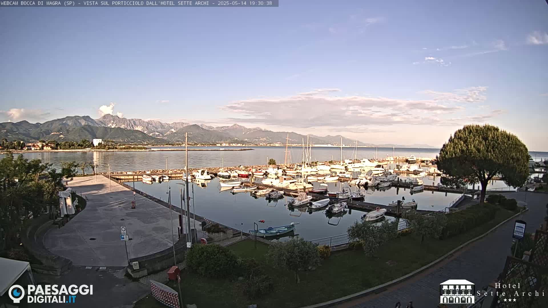 A marina filled with boats is situated near a calm body of water, backed by mountains under a partly cloudy sky.