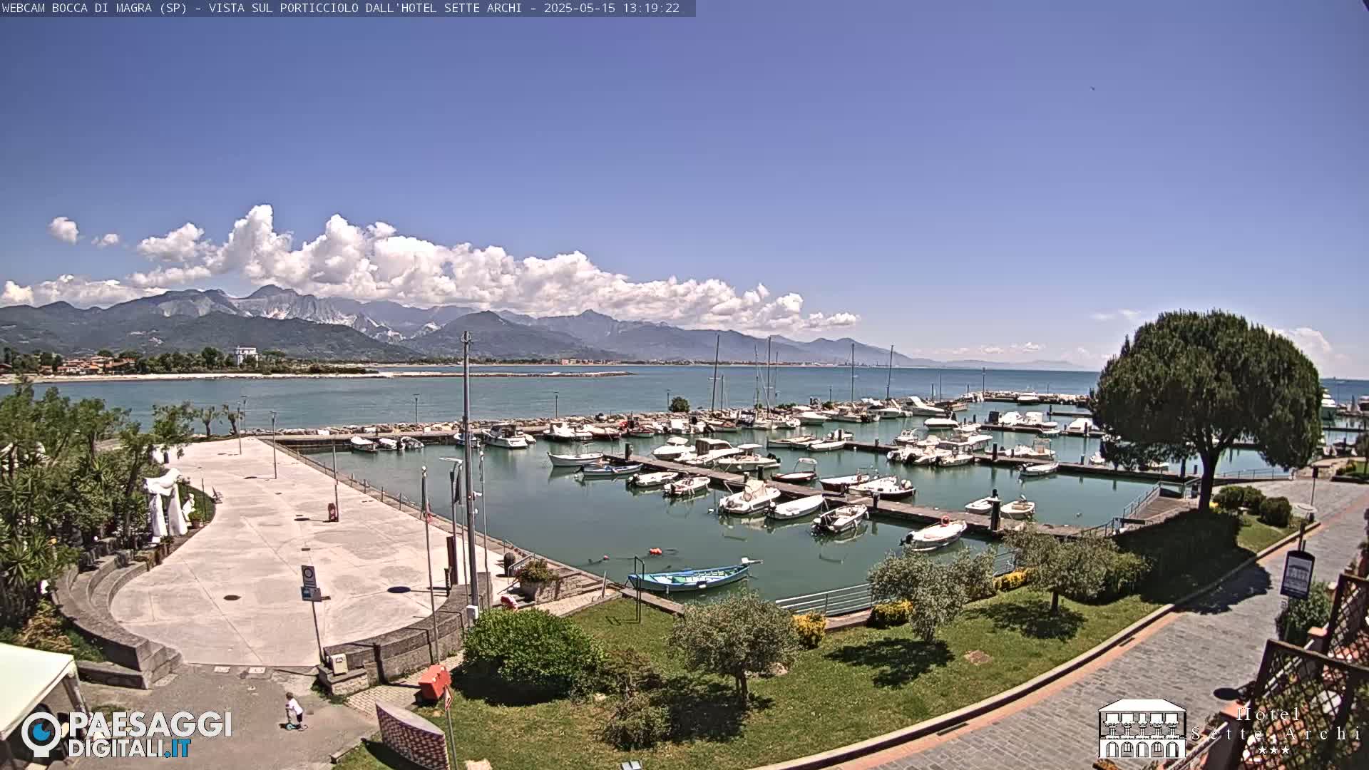 A marina filled with boats is situated next to a paved plaza, all overlooking a calm body of water with mountains in the background under a partly cloudy sky.