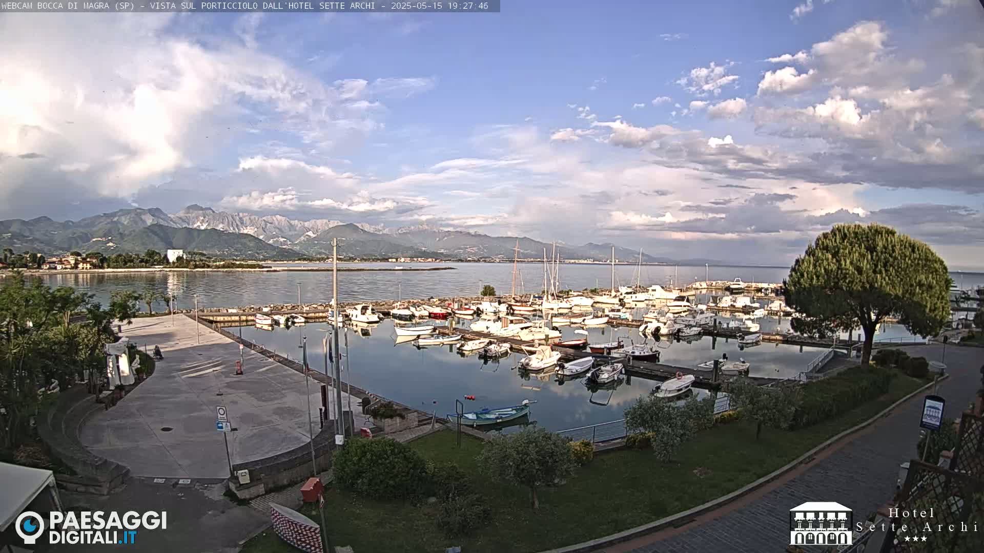 A partly cloudy daytime scene shows a marina filled with numerous boats, a paved area, and green landscaping, all overlooking calm water with mountains in the distance.