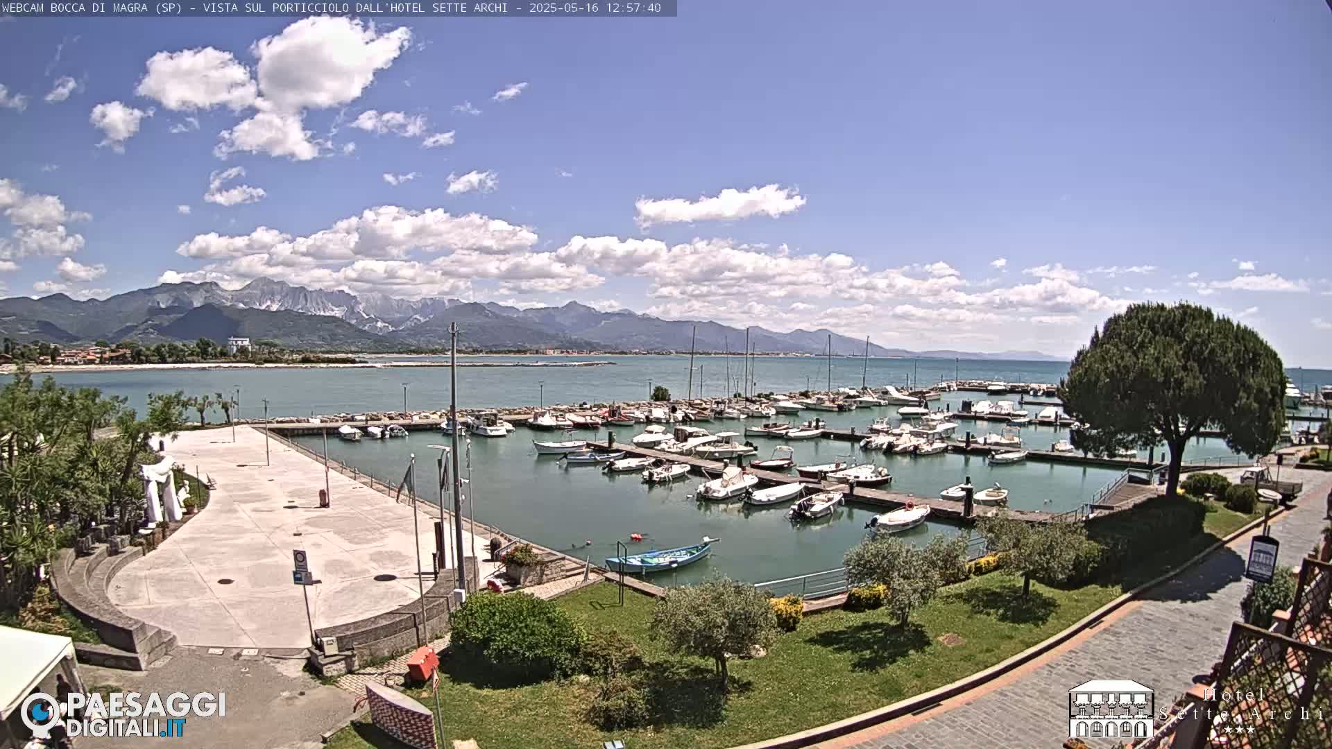 A marina filled with numerous boats is situated beside a paved plaza, all under a partly cloudy sky with mountains visible in the distance.
