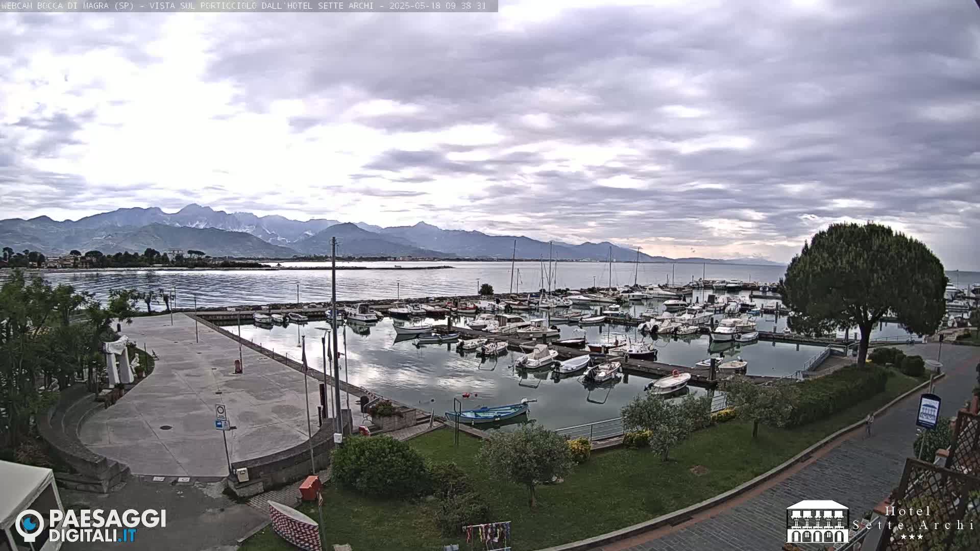 A marina filled with boats is situated next to a paved plaza, overlooking a calm body of water and distant mountains under a cloudy sky.