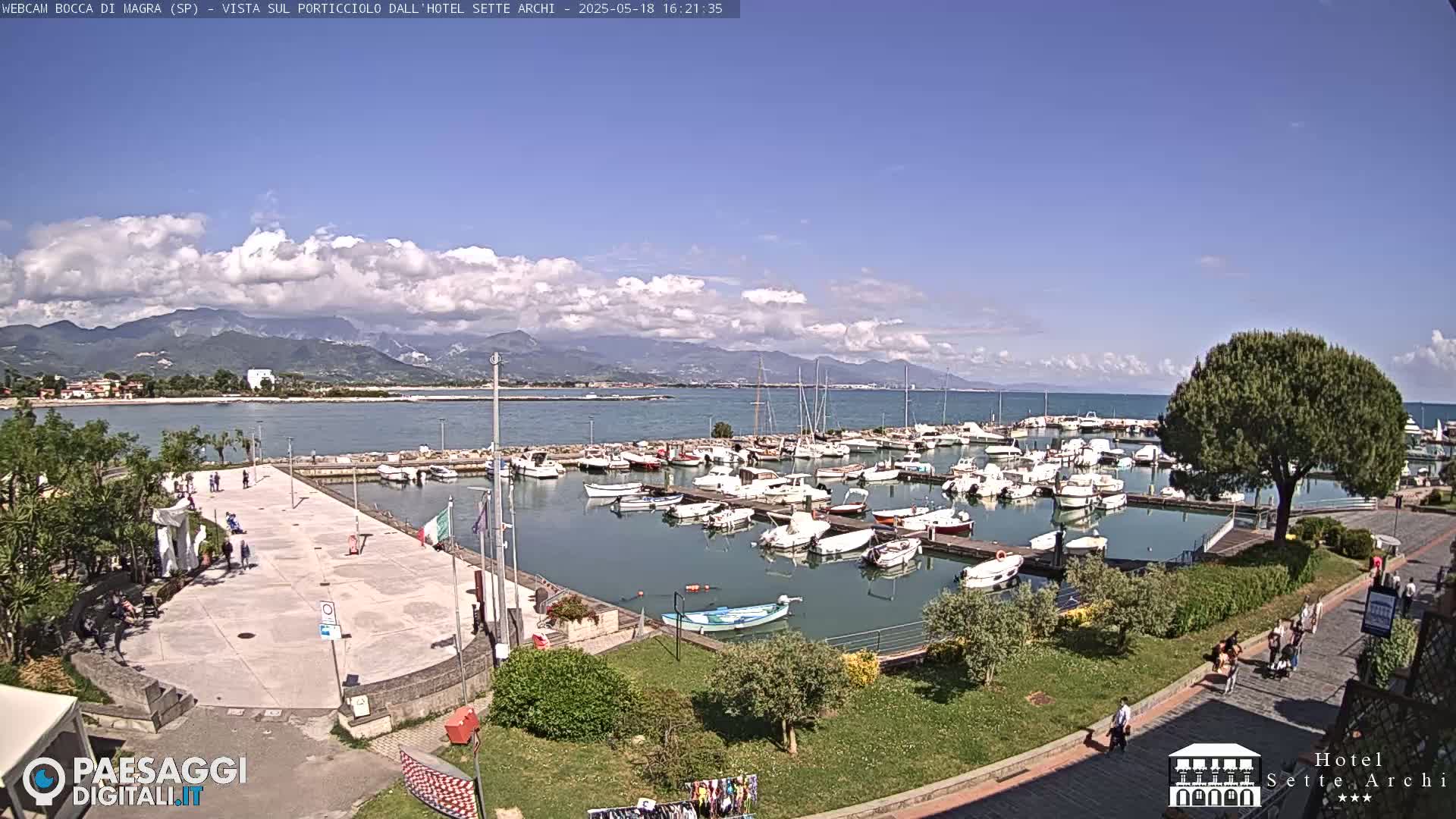 A sunny day with partly cloudy skies overlooks a harbor filled with numerous boats, a paved plaza, and green landscaping, all set against a backdrop of mountains and a calm sea.