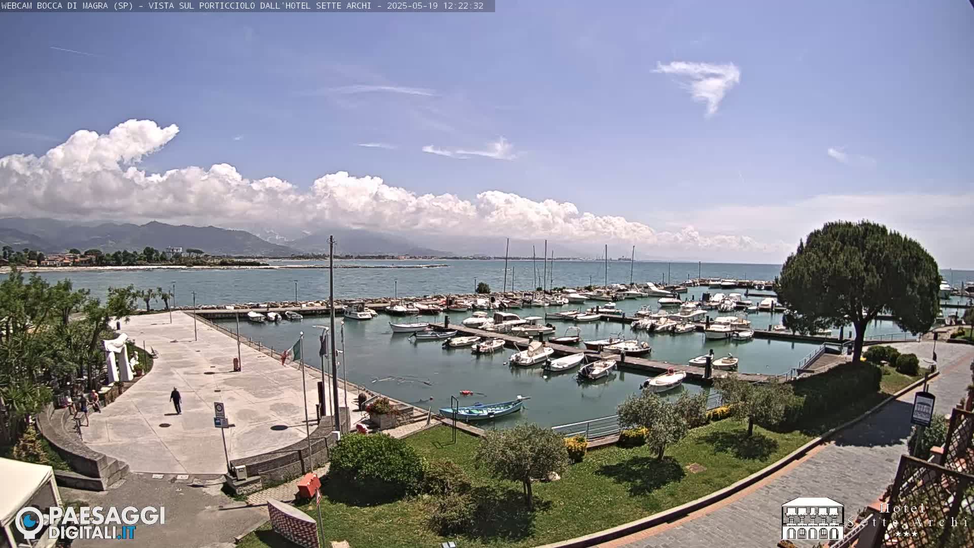 A marina filled with numerous boats is situated next to a paved plaza, all overlooking a calm sea under a partly cloudy sky and a mountain range in the distance.