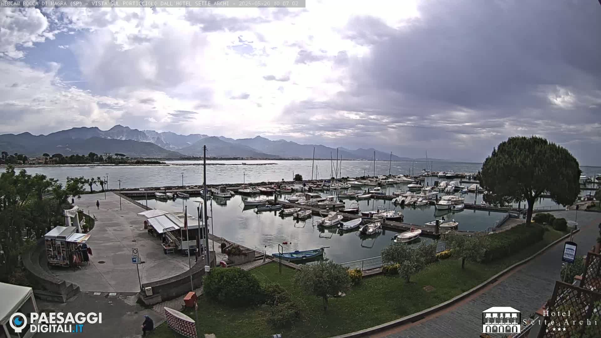 A marina filled with boats is situated near a calm body of water, with mountains in the distance under a cloudy sky.