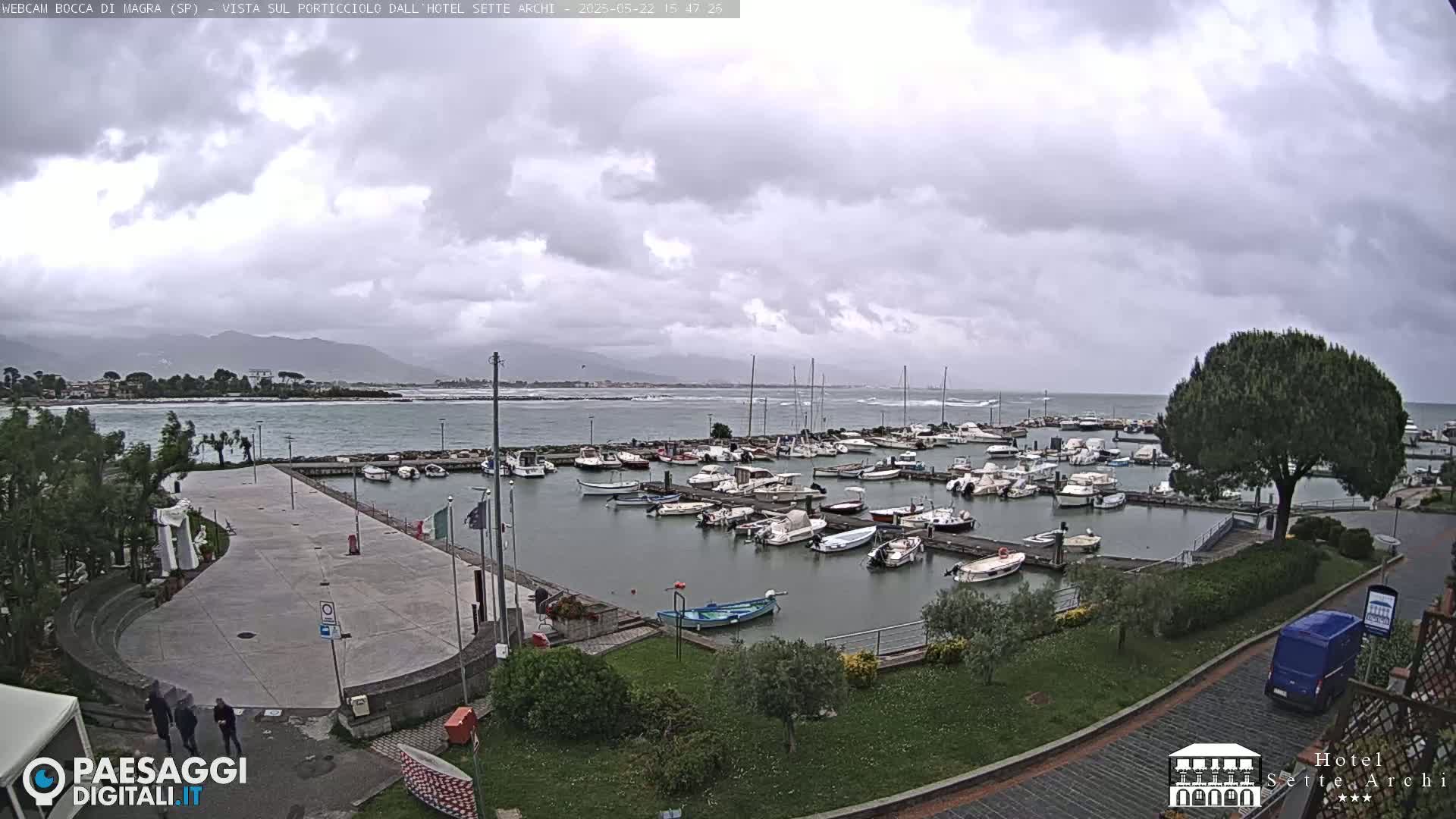 A harbor filled with numerous boats is situated near a paved plaza, all under an overcast sky with distant mountains visible in the background.