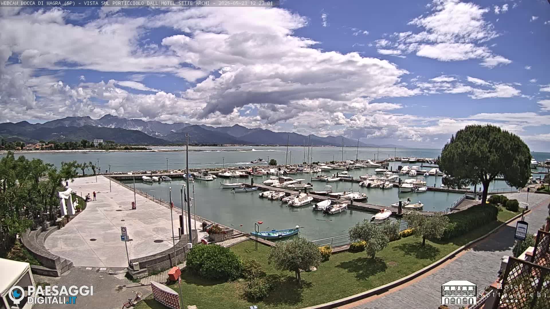 A partly cloudy day shows a marina filled with boats, a paved plaza, and lush green landscaping, all situated against a backdrop of mountains and calm water.