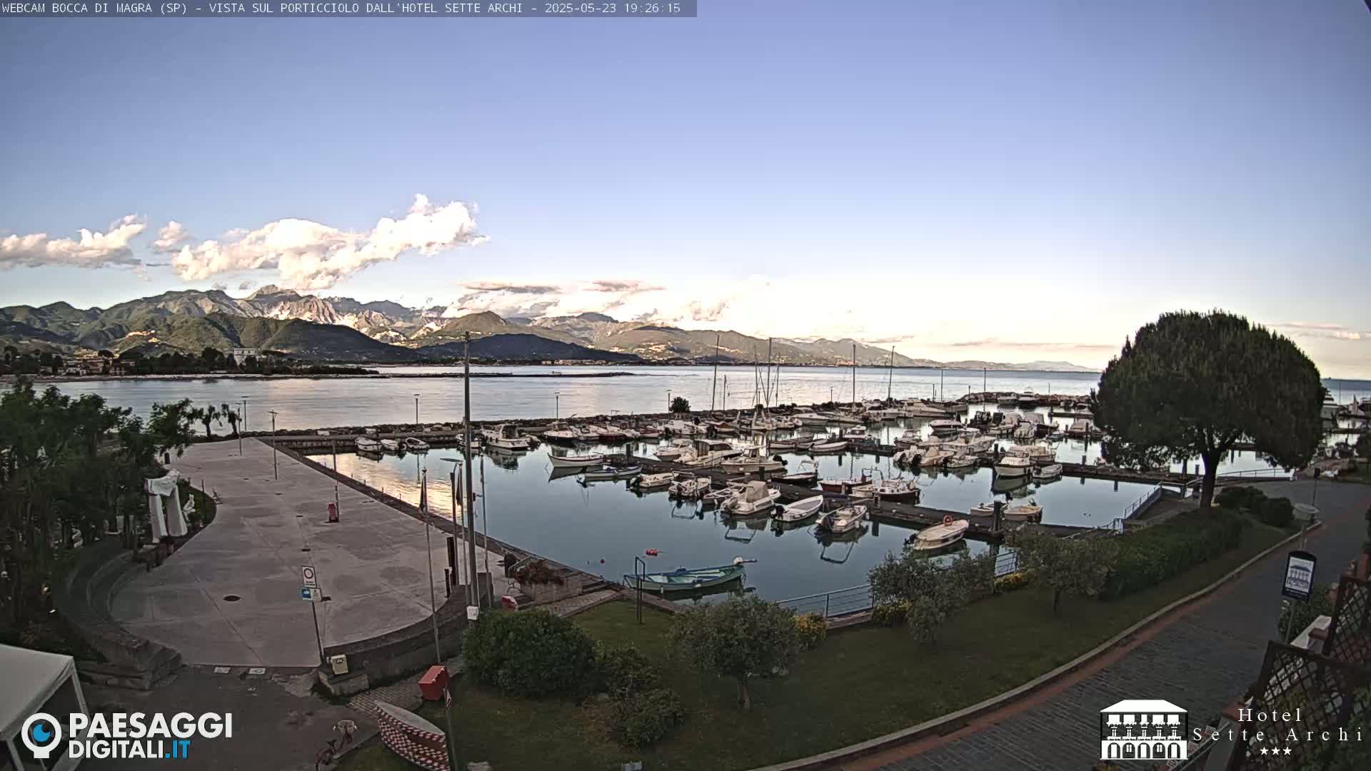 A marina filled with numerous boats is situated near a calm body of water, with mountains in the background under a partly cloudy sky.