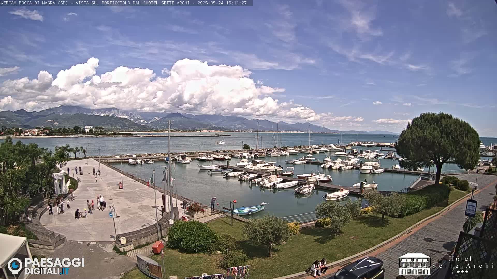 A marina filled with boats is situated near a paved plaza with people walking by, under a partly cloudy sky, with mountains visible in the distance.