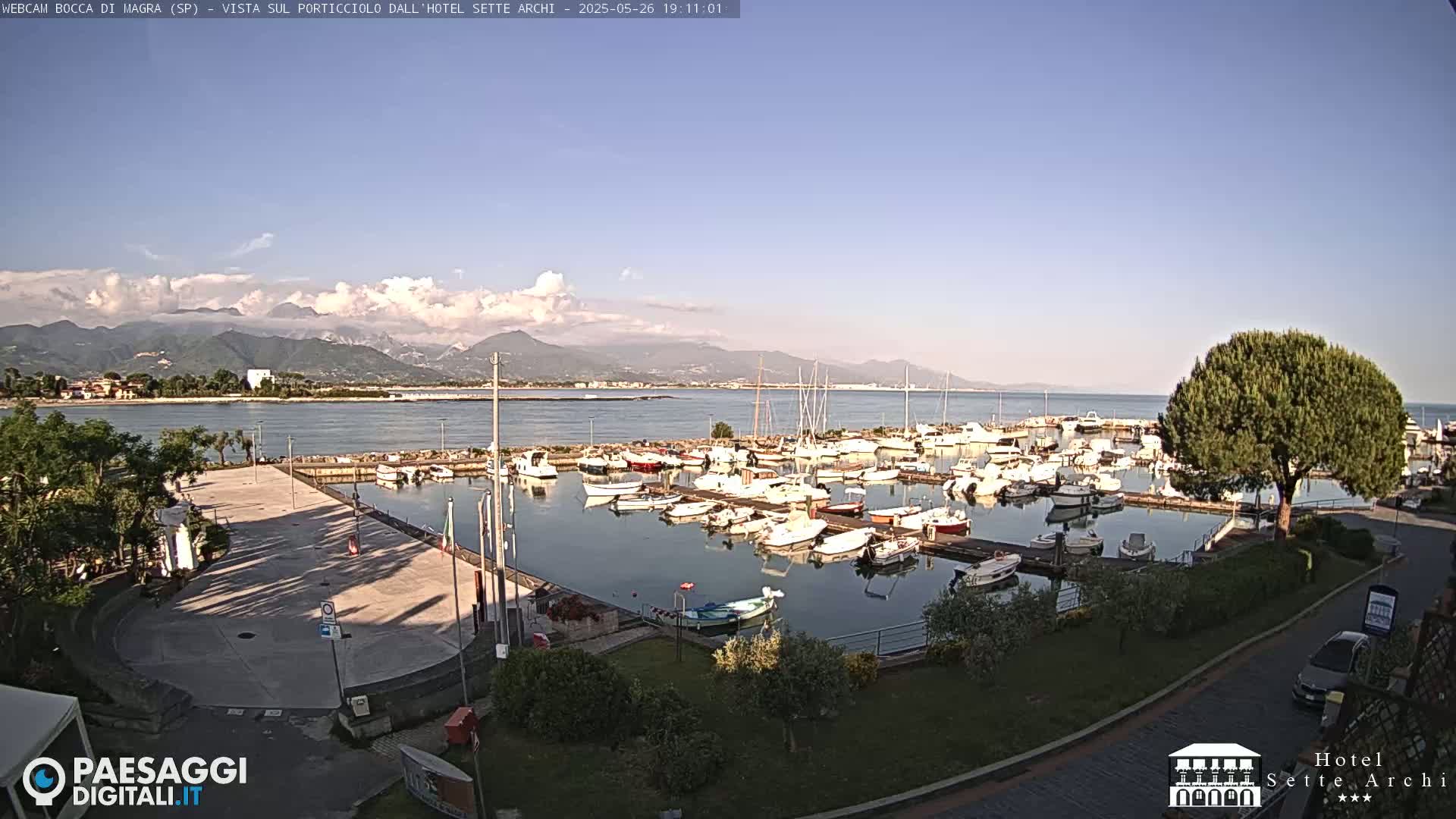 A marina filled with numerous boats is situated near a paved area, with mountains in the background under a mostly clear sky.